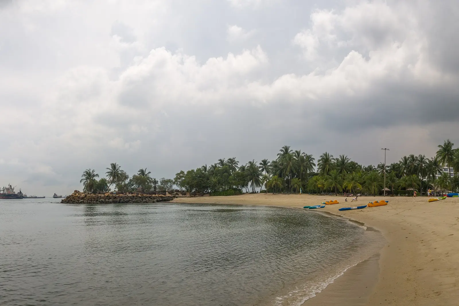 A wide view of Siloso Beach under a grey sky, with sand curving along calm water and small tree-covered islets