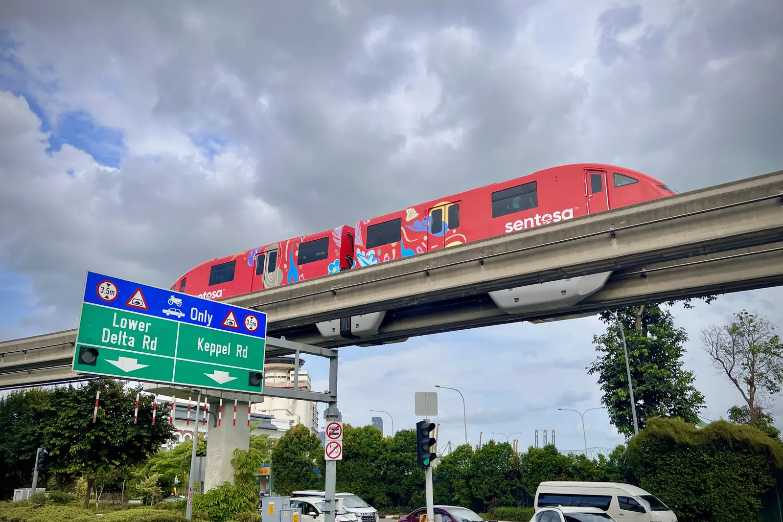 A red Sentosa Express monorail train on its elevated track above a road junction, under a cloudy sky
